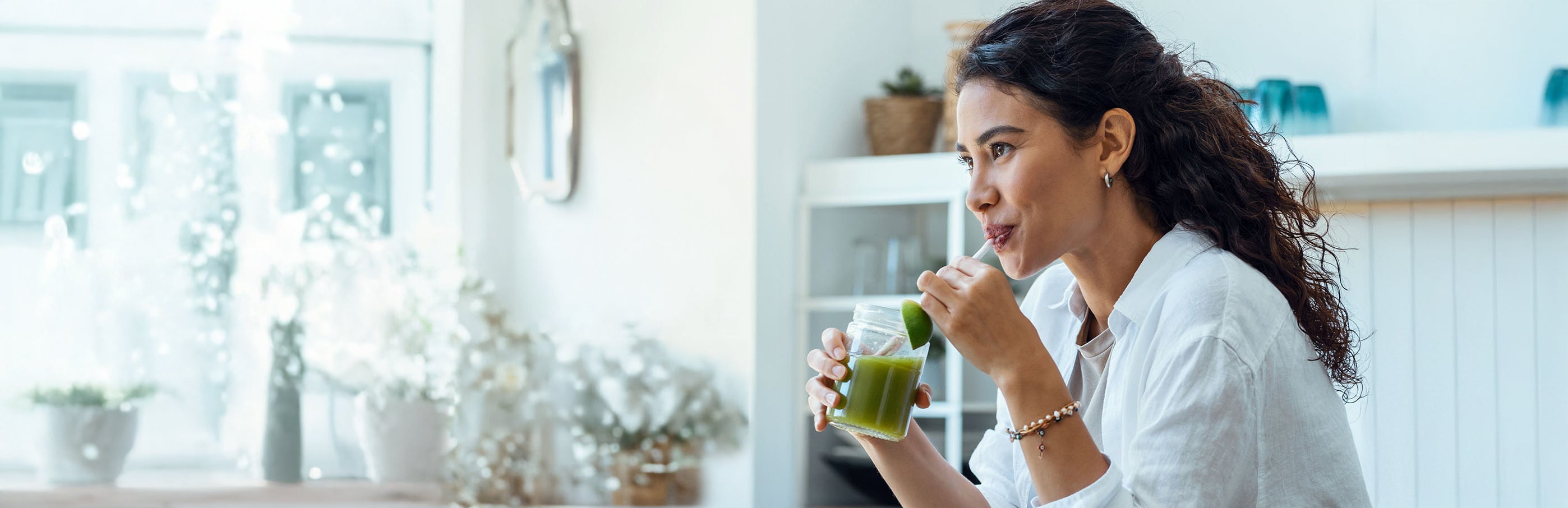 Woman drinking from a green smoothie in a bright kitchen.